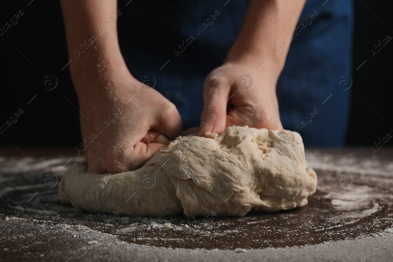 Photo of Making bread. Woman kneading dough at wooden table on dark background, closeup