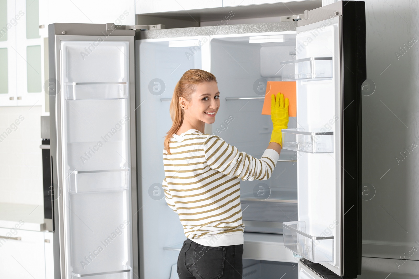 Photo of Woman in rubber gloves cleaning empty refrigerator at home