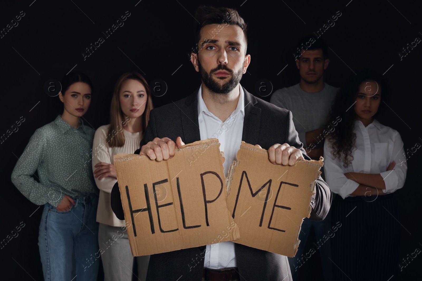 Photo of Unhappy man with HELP ME sign and group of people behind his back on dark background