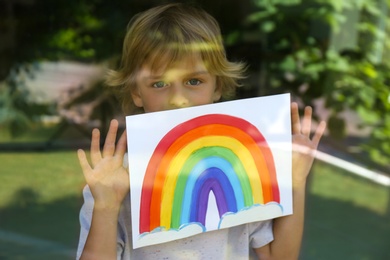 Photo of Little boy with picture of rainbow near window, view from outdoors. Stay at home concept