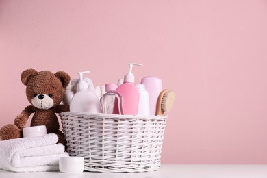 Photo of Baby cosmetic products in wicker basket, bath accessories and knitted toy bear on white table against pink background. Space for text