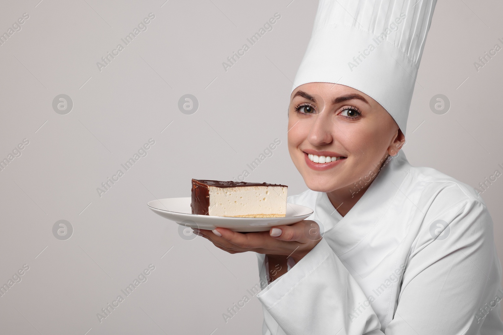 Photo of Happy professional confectioner in uniform holding delicious cheesecake on light grey background. Space for text