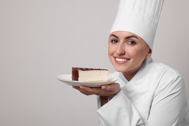 Photo of Happy professional confectioner in uniform holding delicious cheesecake on light grey background. Space for text