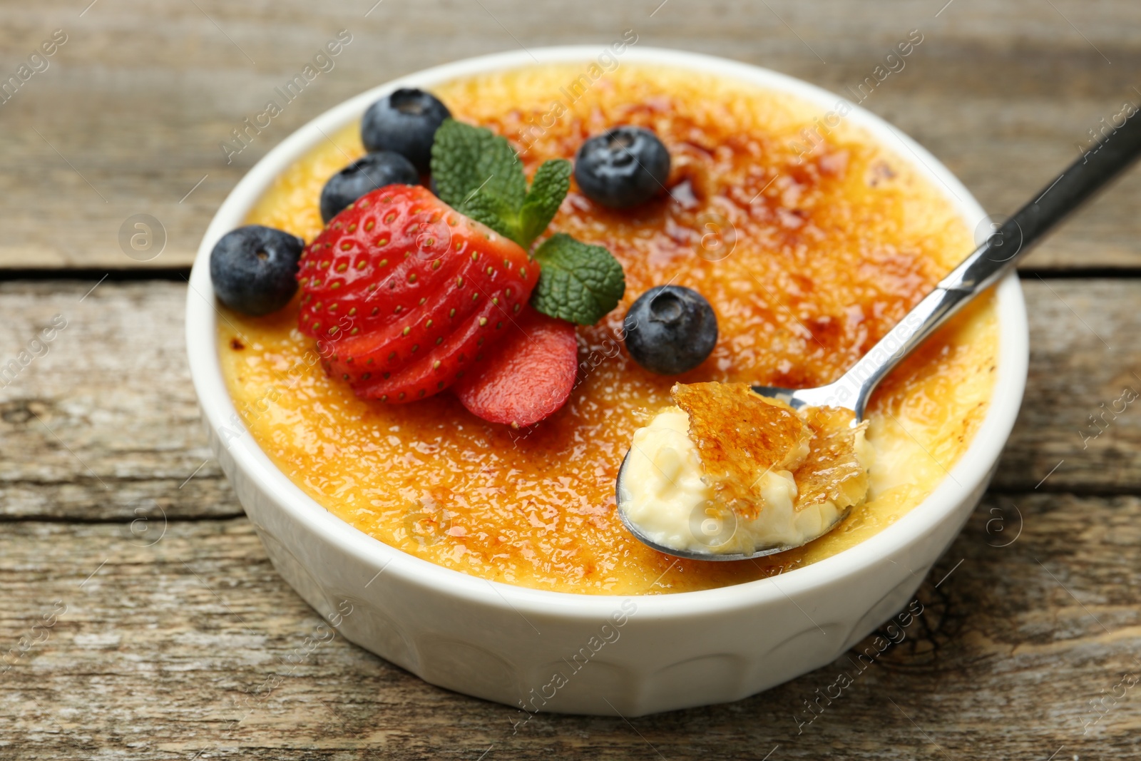 Photo of Delicious creme brulee with berries in bowl and spoon on wooden table, closeup