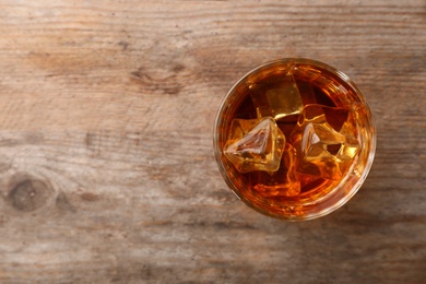 Photo of Golden whiskey in glass with ice cubes on wooden table, top view. Space for text