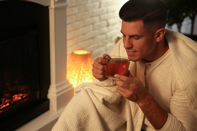 Man covered with white plaid enjoying cup of tea near fireplace