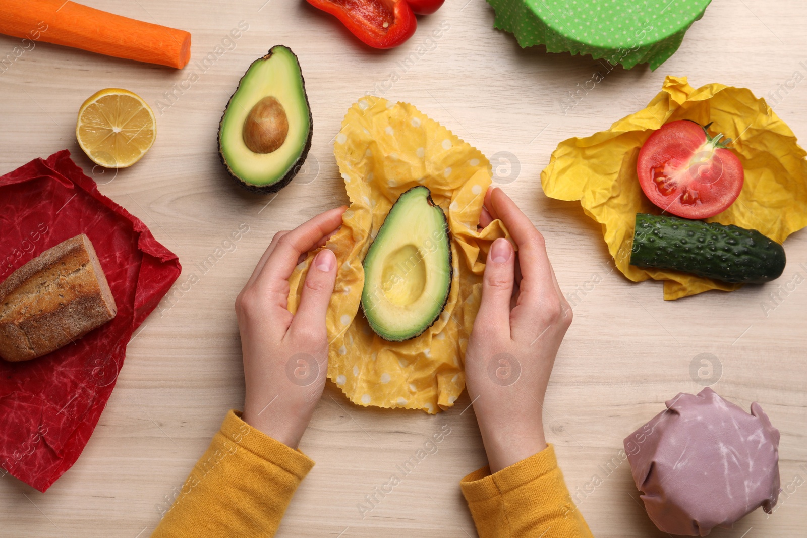 Photo of Woman packing half of fresh avocado into beeswax food wrap at wooden table, top view