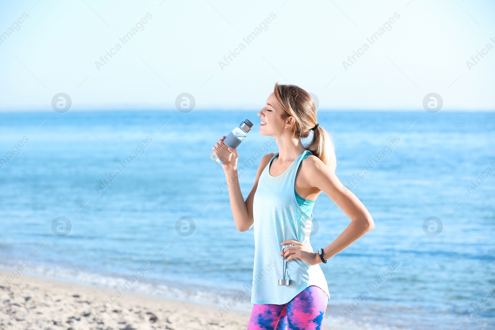 Photo of Young woman drinking water from bottle after fitness exercises on beach in morning