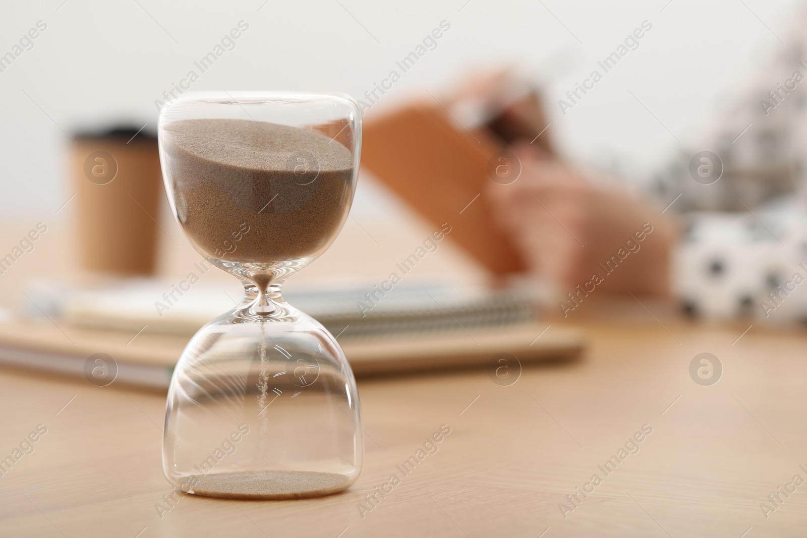 Photo of Hourglass with flowing sand on desk. Woman working indoors, selective focus