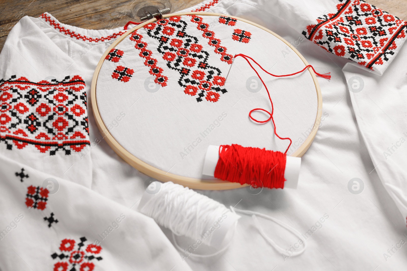 Photo of Shirt with red embroidery design in hoop, needle and thread on table, closeup. National Ukrainian clothes