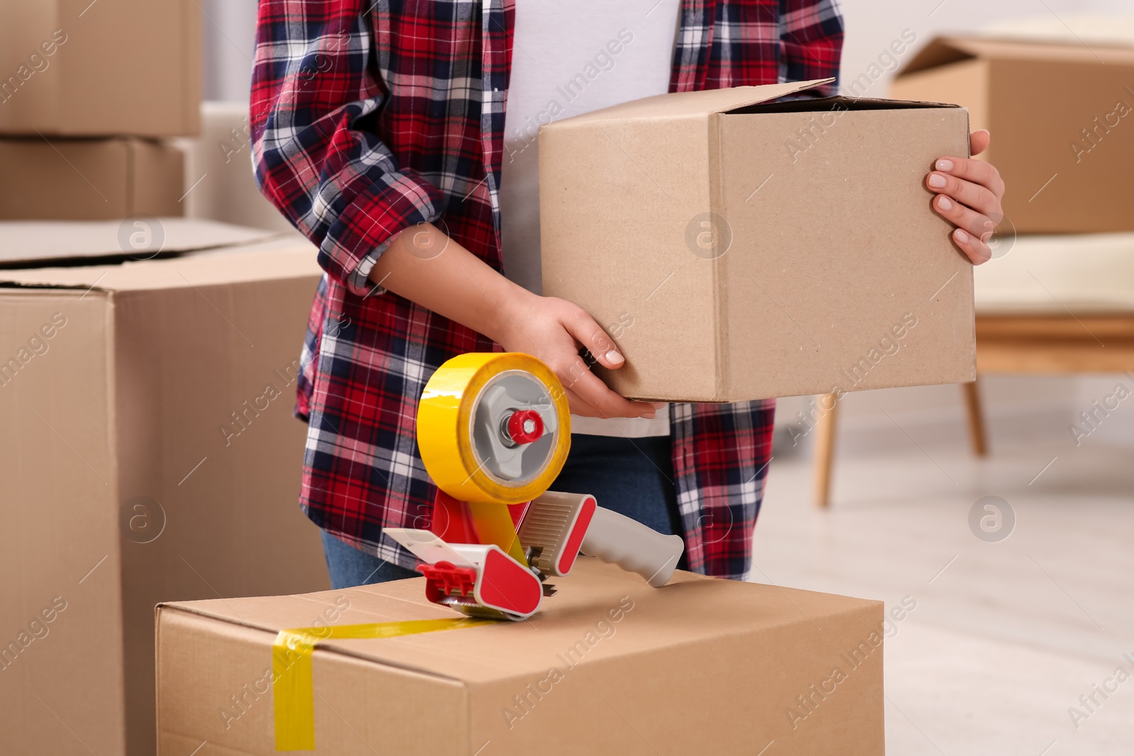 Photo of Woman applying adhesive tape on box with dispenser indoors, closeup