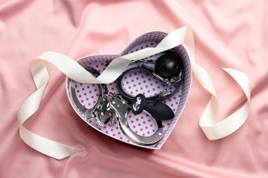 Photo of Heart shaped gift box with different sex toys and ribbon on pink silk fabric, flat lay