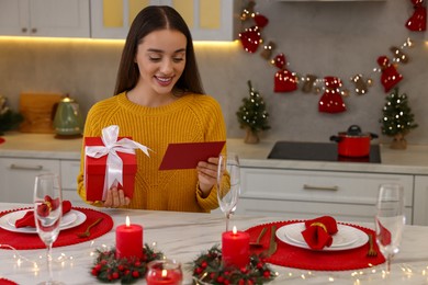 Happy young woman with Christmas gift reading greeting card at table in kitchen