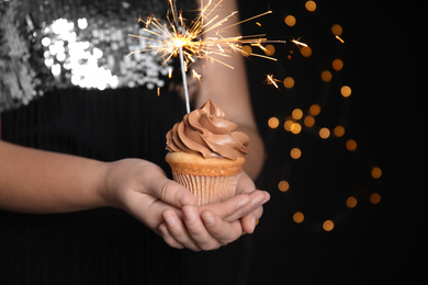 Photo of Woman holding birthday cupcake with sparkler on blurred background, closeup