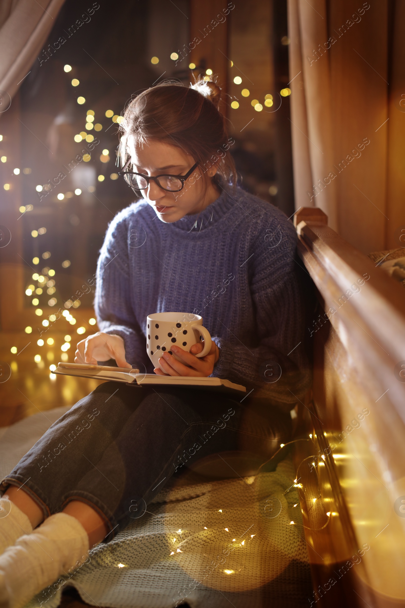 Photo of Woman with cup of hot beverage reading book at home in winter evening