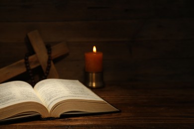 Bible, cross, rosary beads and church candle on wooden table, space for text