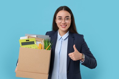 Photo of Happy unemployed woman with box of personal office belongings showing thumbs up on light blue background
