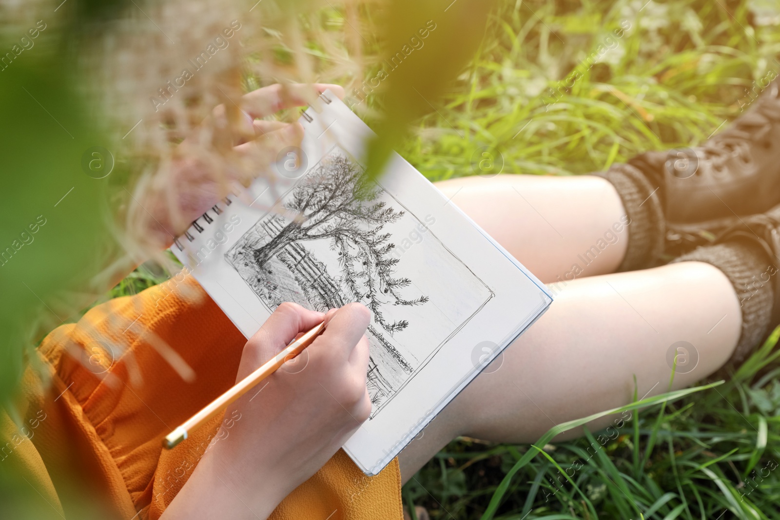 Photo of Young woman drawing with pencil in notepad while sitting on green grass, closeup