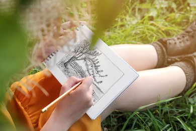 Photo of Young woman drawing with pencil in notepad while sitting on green grass, closeup