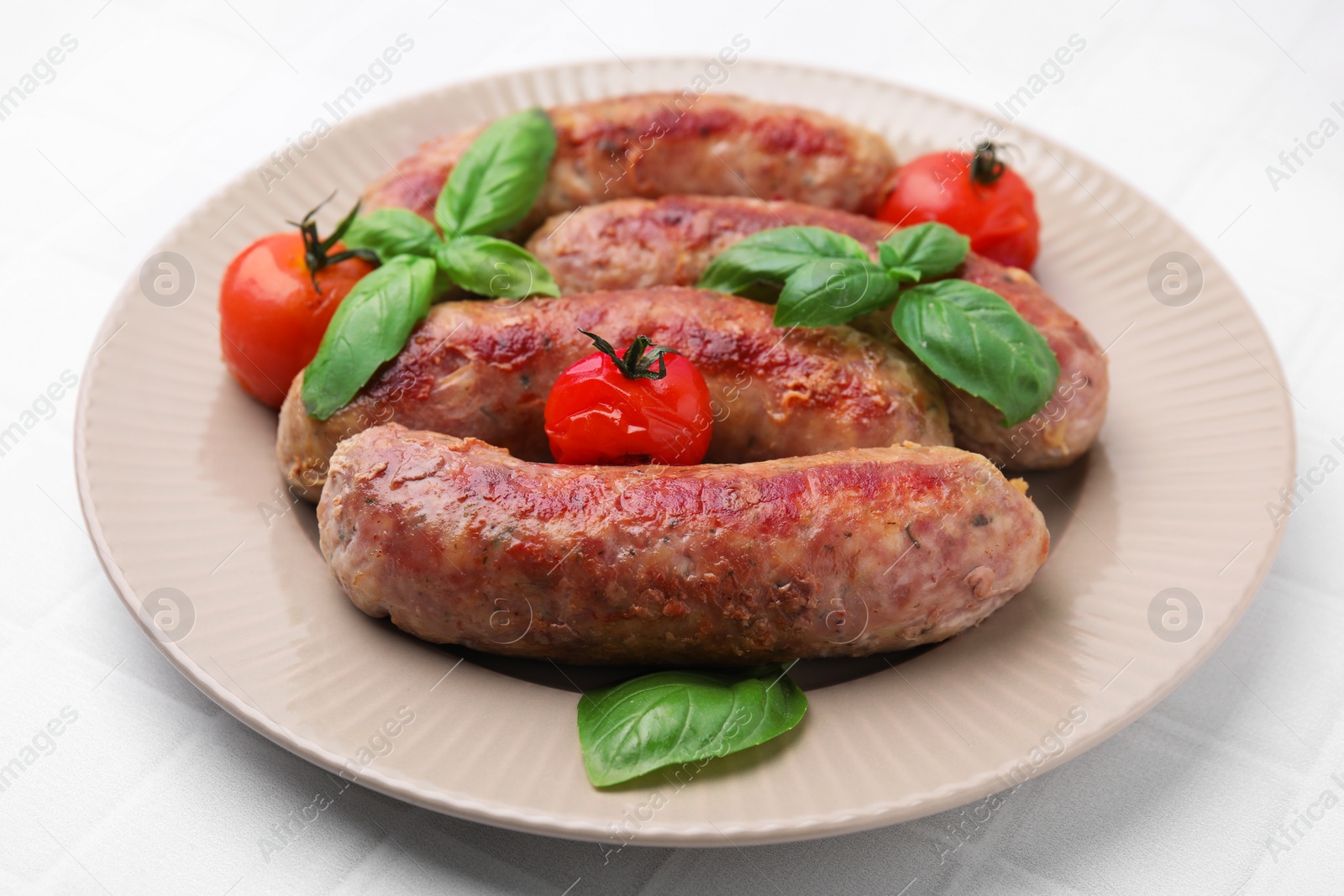 Photo of Plate with tasty homemade sausages, basil leaves and tomatoes on white tiled table, closeup