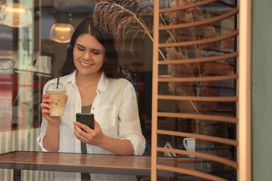 Photo of Young beautiful woman with smartphone drinking coffee in cafe, view through window glass