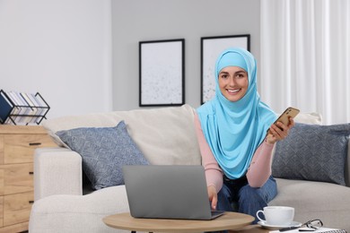 Muslim woman with smartphone using laptop at couch in room. Space for text