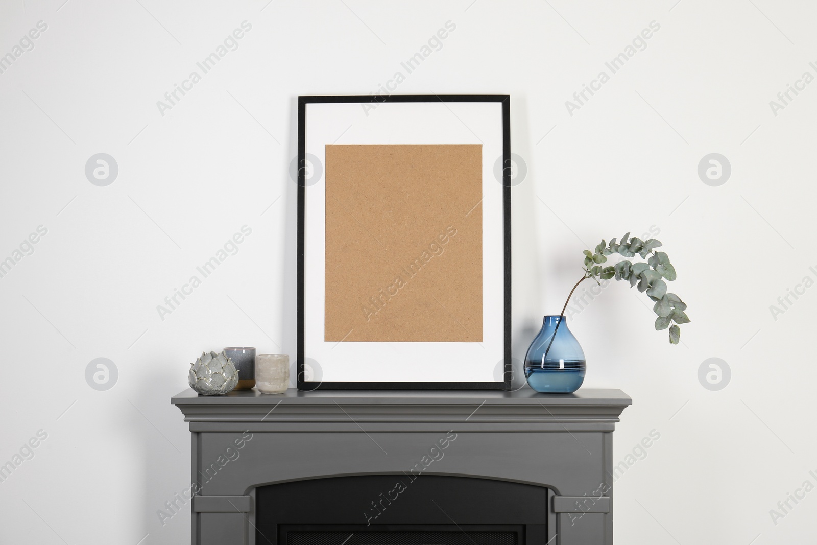Photo of Empty frame, candles and eucalyptus branch in vase on fireplace near white wall indoors. Interior design