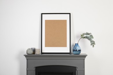 Empty frame, candles and eucalyptus branch in vase on fireplace near white wall indoors. Interior design