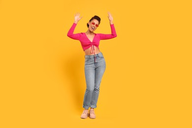 Happy young woman in stylish sunglasses dancing on orange background