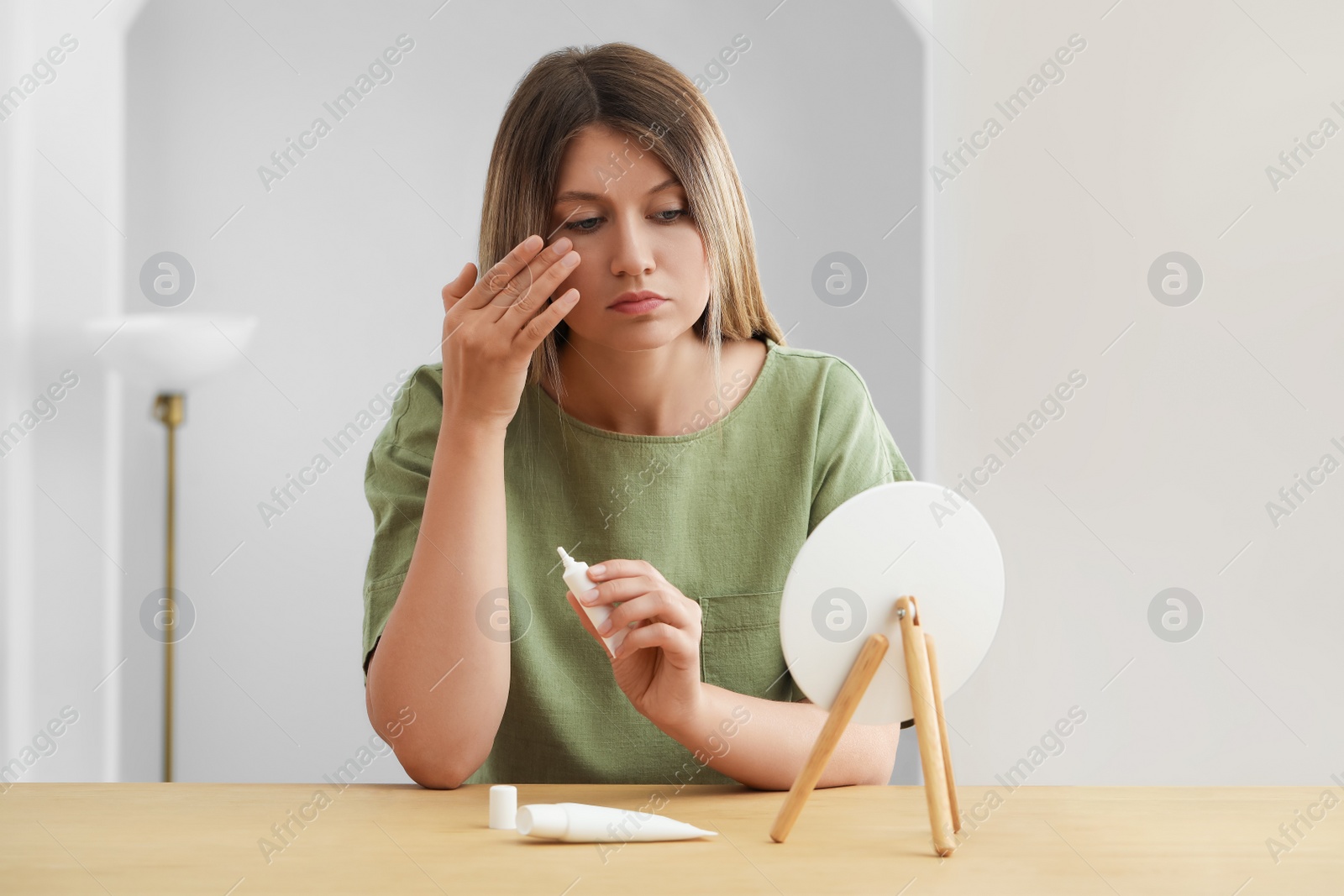 Photo of Sleep deprived young woman covering up dark circles with concealer near mirror at home