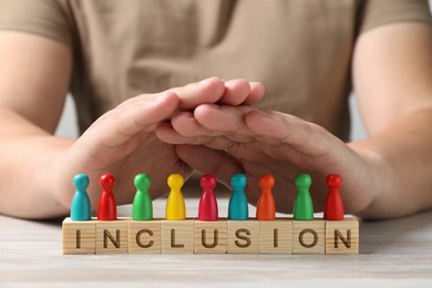 Photo of Woman protecting colorful pawns and cubes with word Inclusion at wooden table, closeup