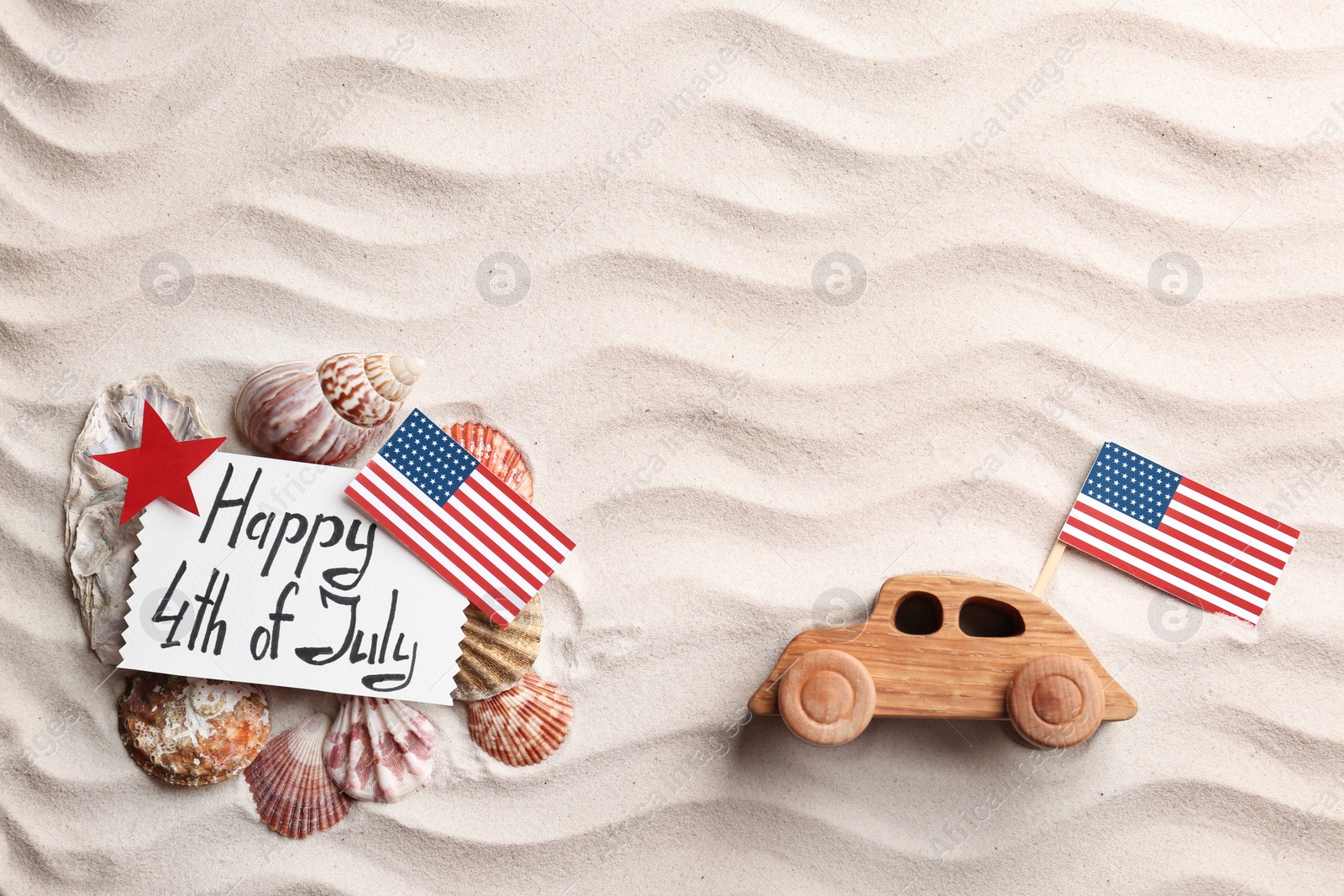 Photo of Flat lay composition with greeting card, USA flags and wooden car on sand. Happy Independence Day