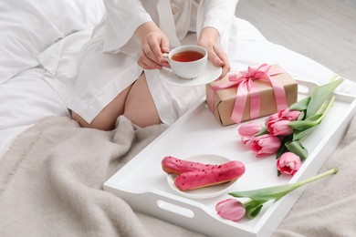 Photo of Tasty breakfast served in bed. Woman with tea, eclairs, gift box and flowers at home, closeup