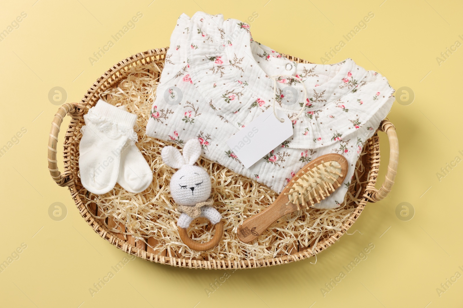 Photo of Different baby accessories, clothes and blank card in wicker basket on yellow background, top view