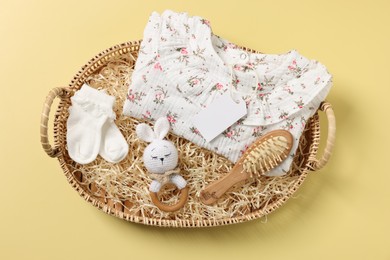 Different baby accessories, clothes and blank card in wicker basket on yellow background, top view