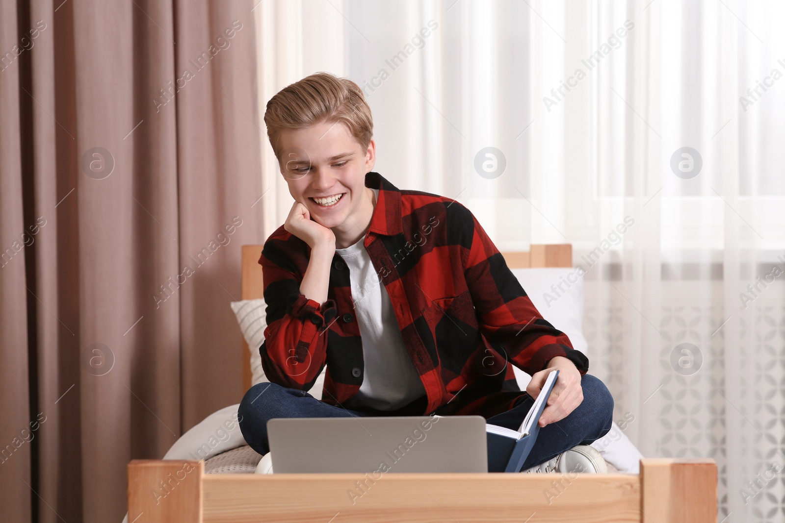 Photo of Online learning. Smiling teenage boy with book looking on laptop at home