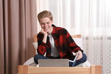 Photo of Online learning. Smiling teenage boy with book looking on laptop at home