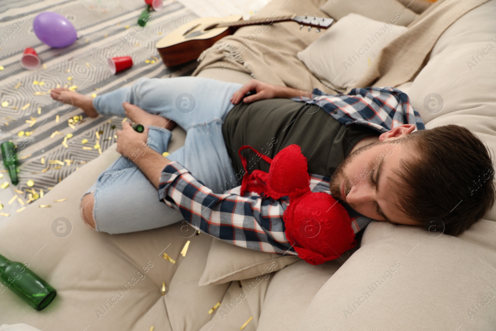 Photo of Young man with red bra sleeping on sofa in messy room after party