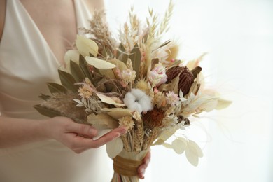 Photo of Bride holding beautiful dried flower bouquet near window at home, closeup