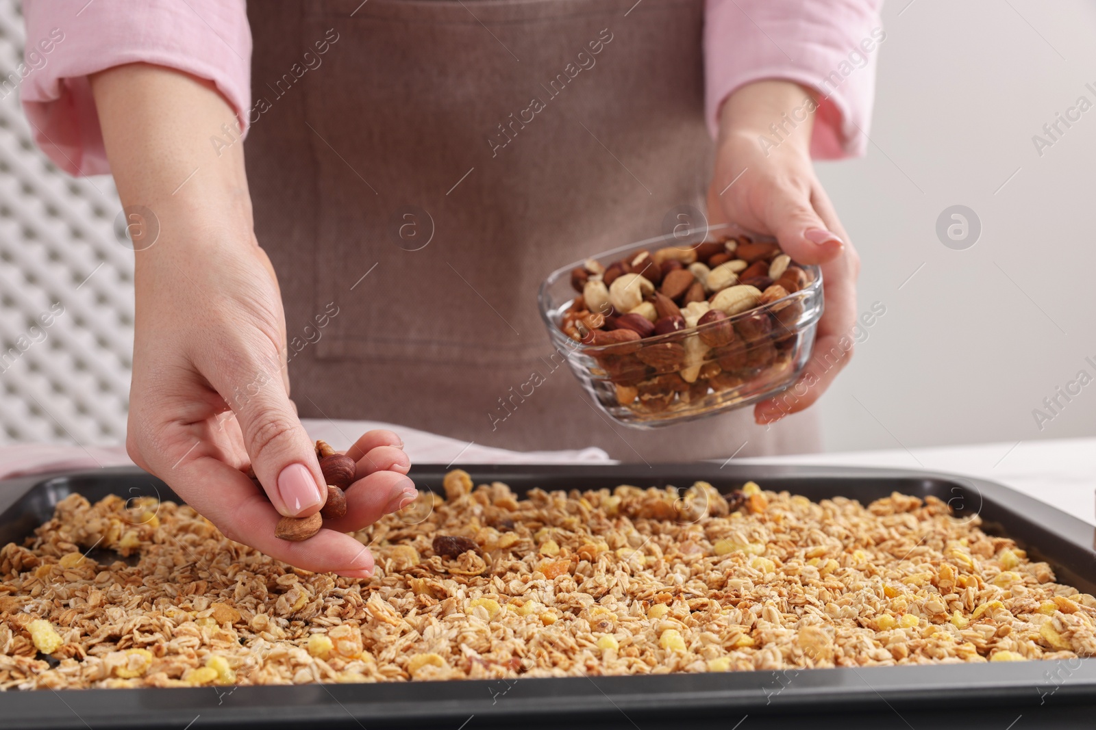 Photo of Making granola. Woman adding nuts onto baking tray with mixture of oat flakes and other ingredients at table in kitchen, closeup