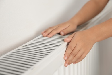 Photo of Child warming hands on heating radiator near white wall, closeup