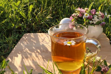 Cup of aromatic herbal tea, pestle and ceramic mortar with different wildflowers on green grass outdoors. Space for text