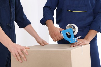 Photo of Male movers packing box with adhesive tape indoors, closeup