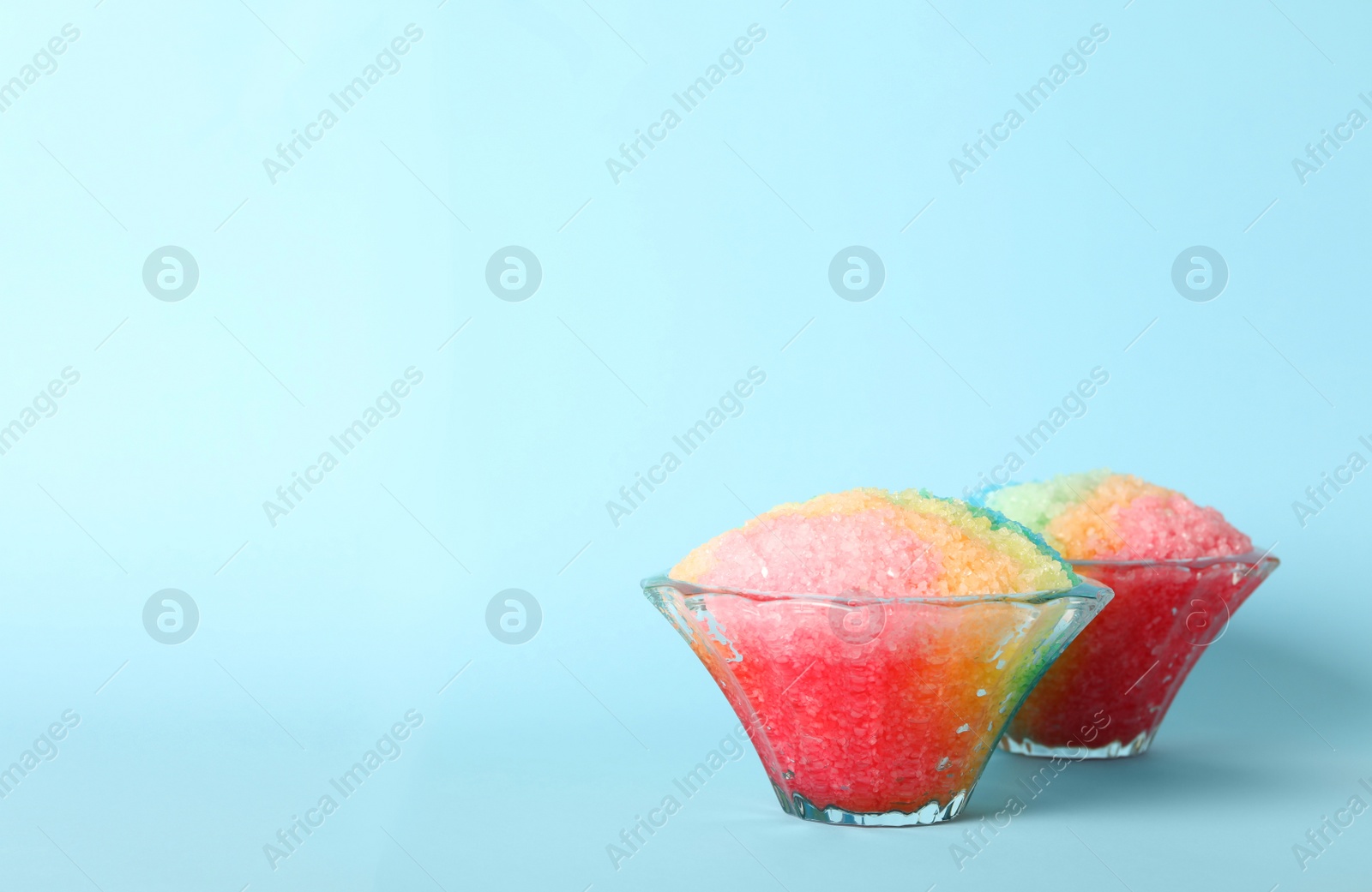 Photo of Rainbow shaving ice in glass dessert bowls on light blue background, space for text