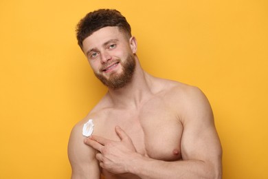 Handsome man applying moisturizing cream onto his shoulder on orange background