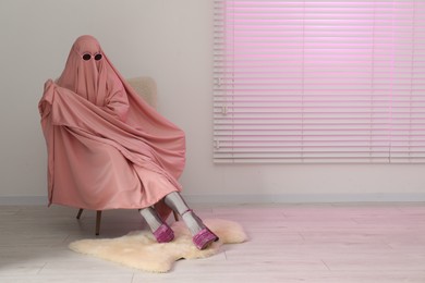Photo of Glamorous ghost. Woman in pink sheet and high heel shoes on armchair indoors, space for text
