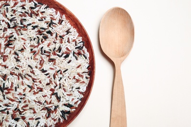 Mixed brown and other types of rice in wooden plate near spoon on light background, top view