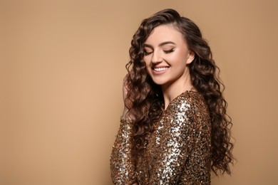 Beautiful young woman with long curly brown hair in golden sequin dress on beige background, space for text