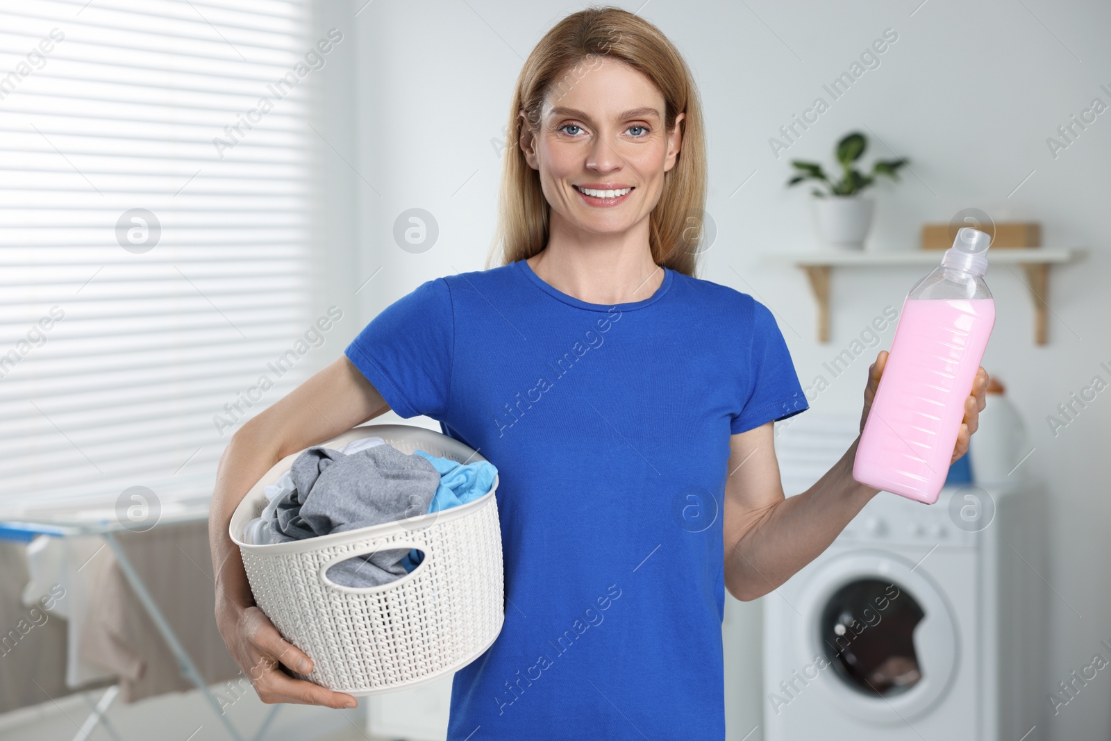 Photo of Woman holding fabric softener and basket with dirty clothes in bathroom