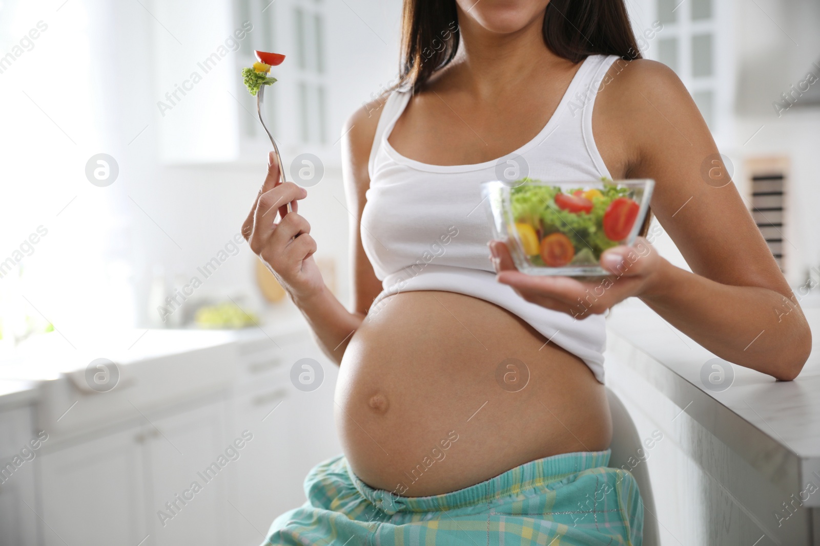 Photo of Young pregnant woman with bowl of vegetable salad at table in kitchen, closeup. Taking care of baby health